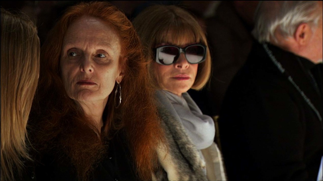 Two women in a dark setting, one wearing large oversized sunglasses and a grey sweater, the other with reddish hair looking concerned.