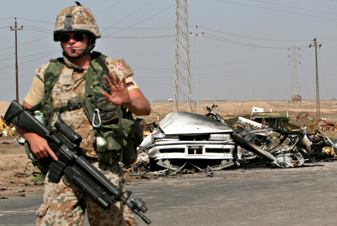 Armed soldier in camouflage uniform standing near destroyed vehicle wreckage in desert landscape.