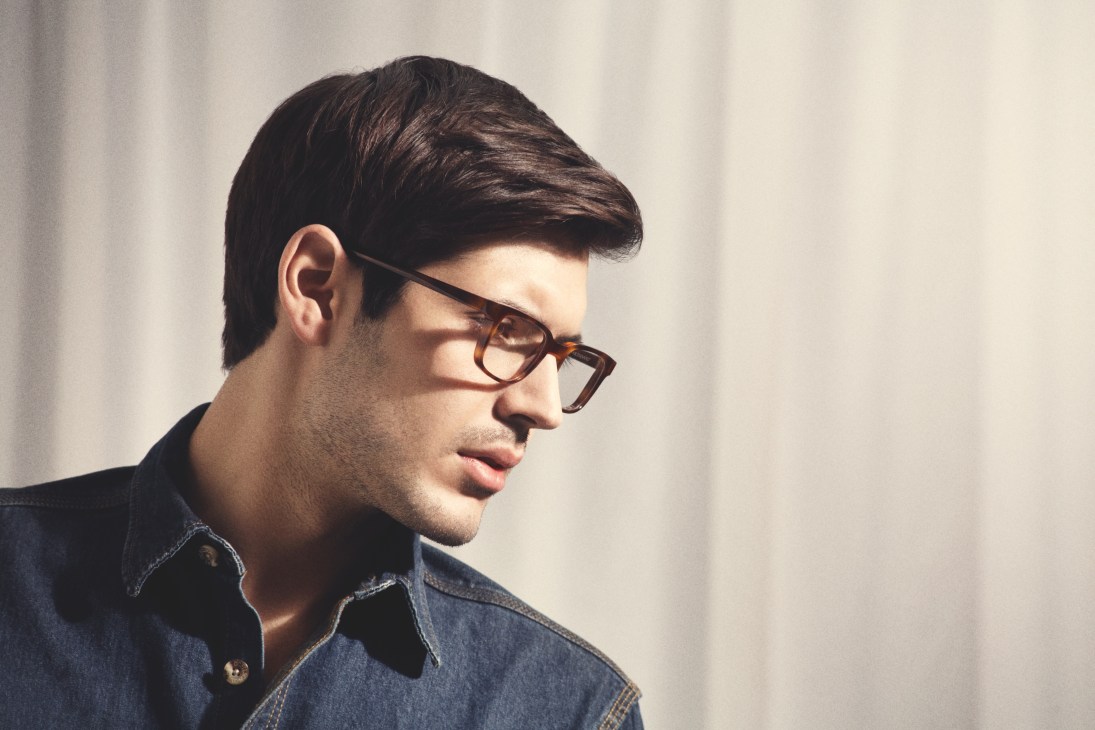 Man wearing tortoiseshell-framed glasses and denim shirt in profile against neutral background.