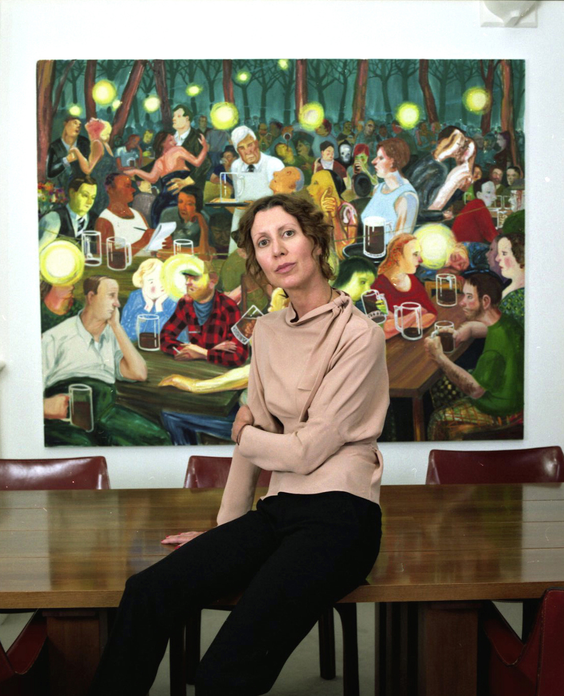 Valeria Napoleone sits on a bench in front of a colorful painting of people at an outdoor gathering.