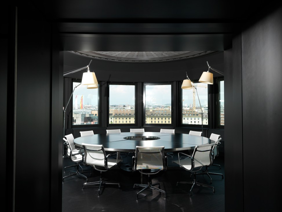 Circular boardroom with round dark table, white Eames chairs, pendant lights, and panoramic windows overlooking Geneva cityscape.