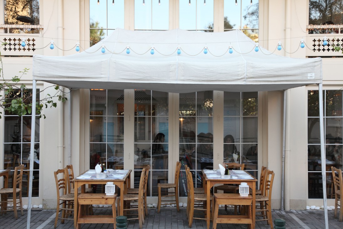 Outdoor seating area at Sardelles restaurant with wooden chairs under white awning and large windows.