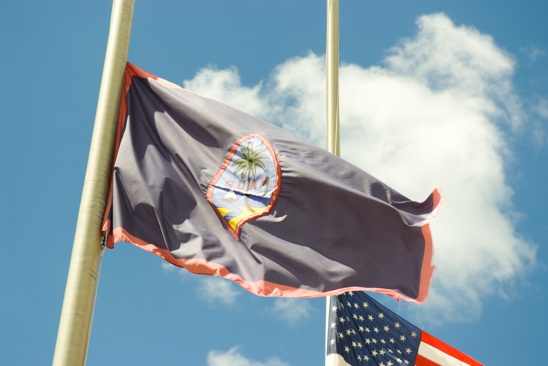 Guam's flag featuring a coconut palm tree and beach design, flying alongside the American flag against a blue sky with white clouds.