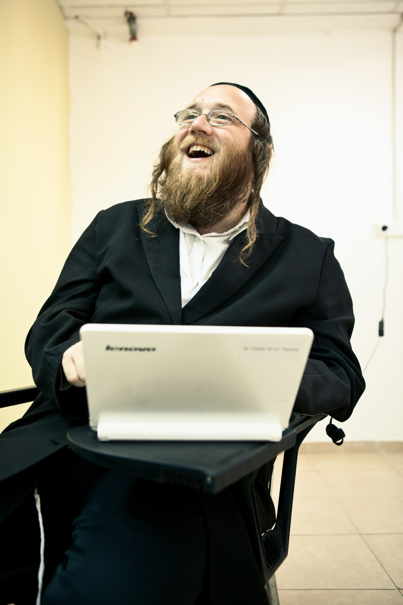 Orthodox Jewish man with beard and glasses holding a laptop computer, smiling indoors.