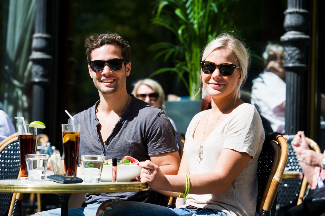 Two people wearing sunglasses sit at an outdoor café table with drinks and food.