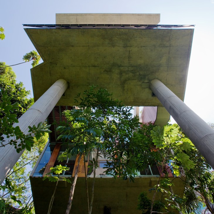Modern concrete house structure supported by two white cylindrical pillars, surrounded by forest vegetation.