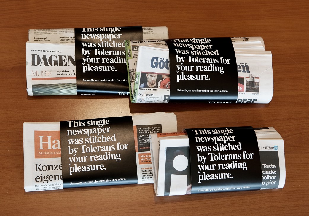 Four European compact newspapers displayed on a wooden surface, each wrapped with black bands reading 'This single newspaper was stitched by Tolerans for your reading pleasure.'