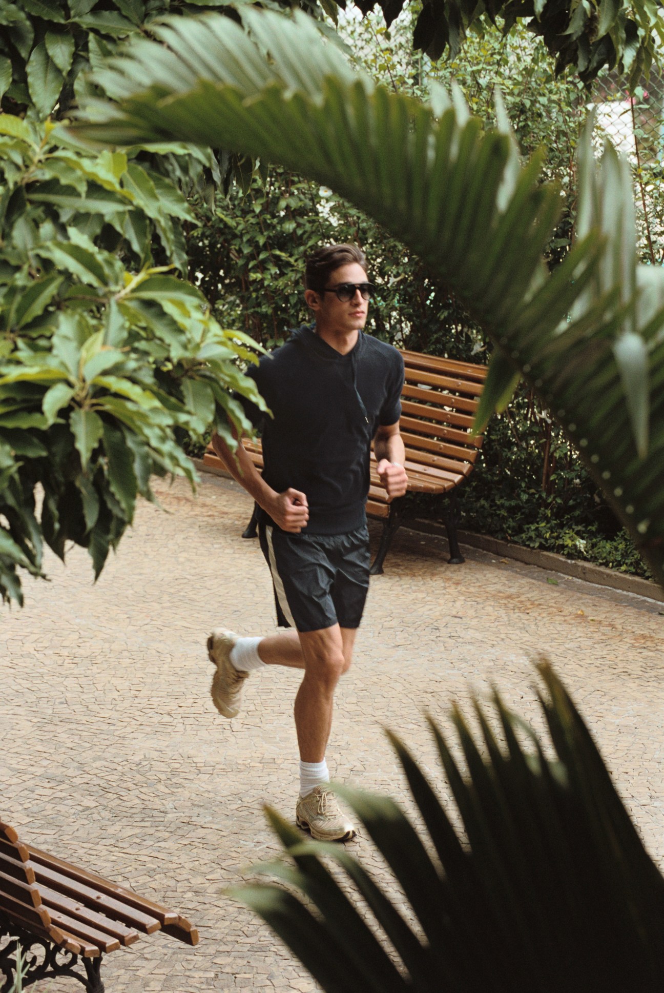 Man jogging through lush garden pathway wearing hooded top by United Arrows, Moncler swimming trunks, and Mizuno shoes at Clube Hebraica.