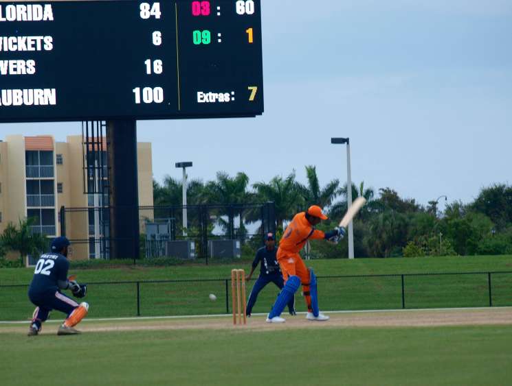 Cricket match in Florida with scoreboard showing Auburn batting, batter in orange uniform at crease.