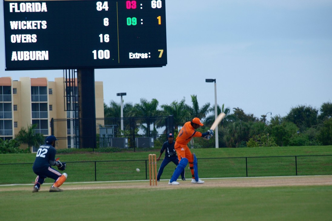 Cricket match in Florida with scoreboard showing Auburn batting, batter in orange uniform at crease.