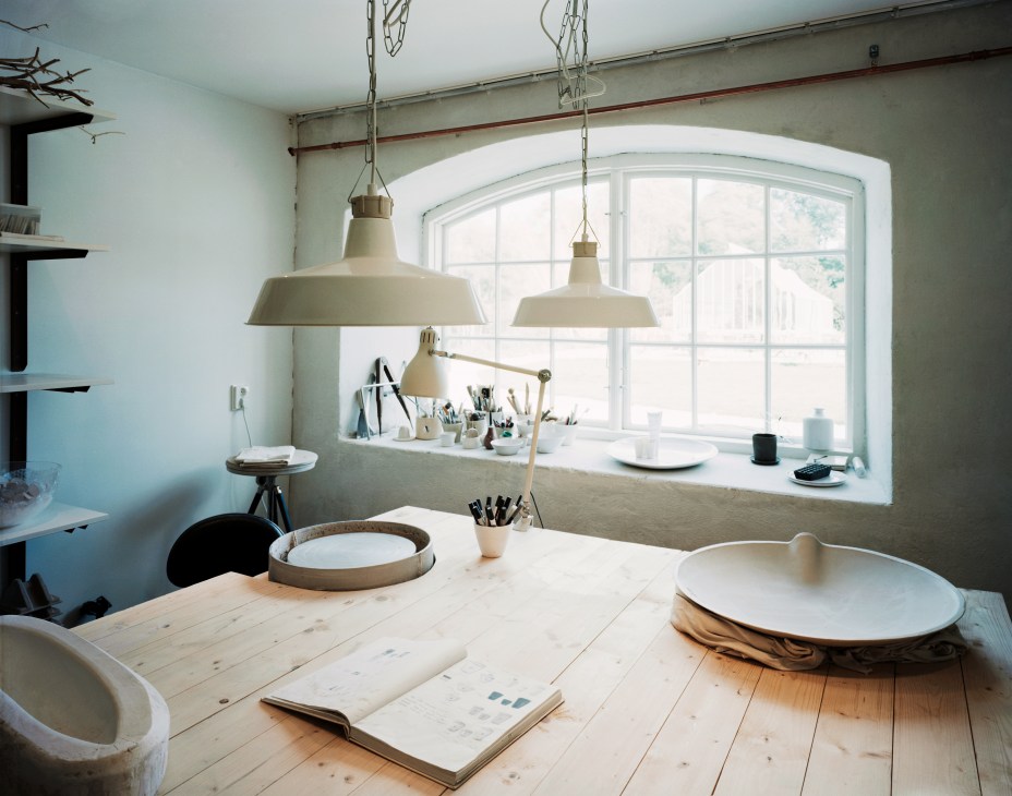 A bright artist's studio workspace with a wooden table, pendant lamps, ceramic bowls, and art supplies.