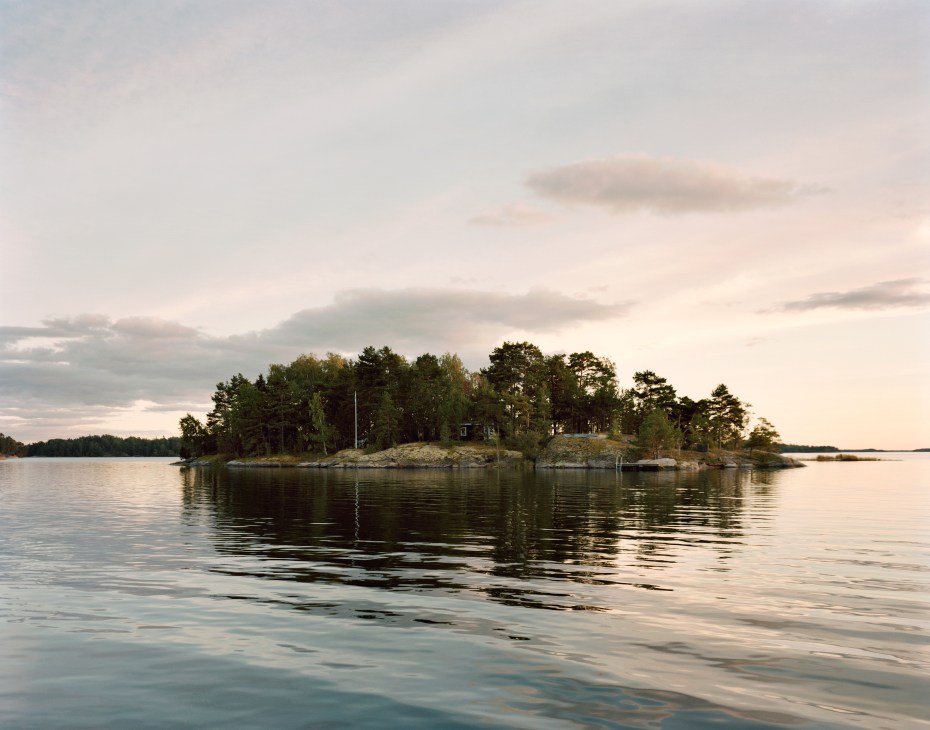 A small island with pine and birch trees and a single-storey house surrounded by calm water in the Swedish archipelago at dusk.