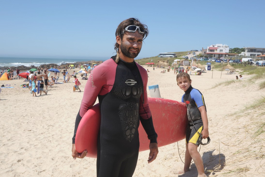 Surf instructor in wetsuit carrying red surfboard on La Pedrera beach with students.