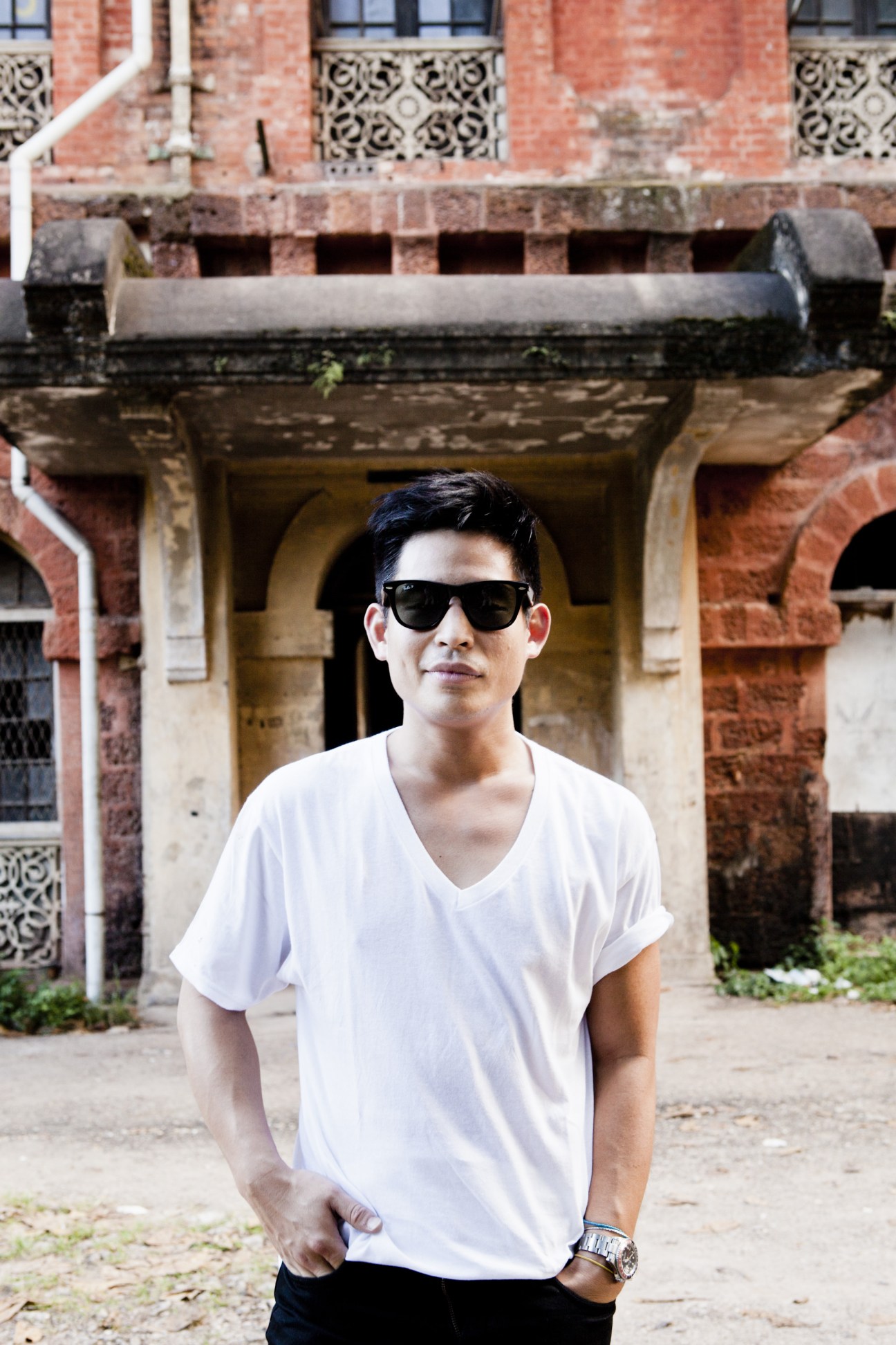 Young man in white shirt and sunglasses standing in front of deteriorating colonial brick building with decorative balcony railings.
