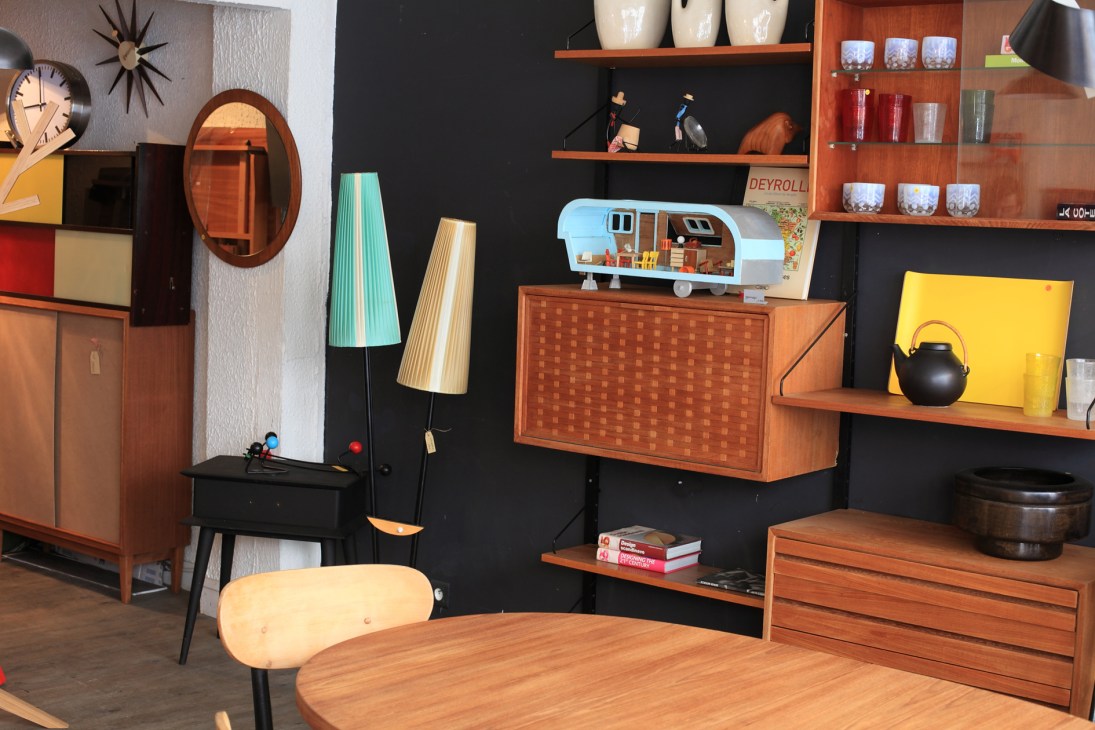 Vintage mid-century furniture display including wooden cabinets, shelving, lamps, and a decorative caravan model.
