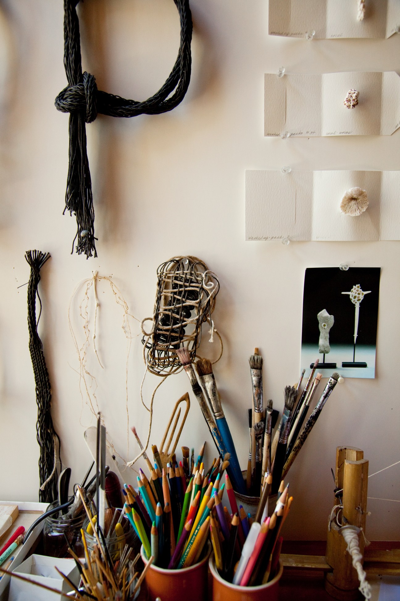 Artist's studio wall displaying colorful pencils, paintbrushes, black thread, woven samples, and small art objects.