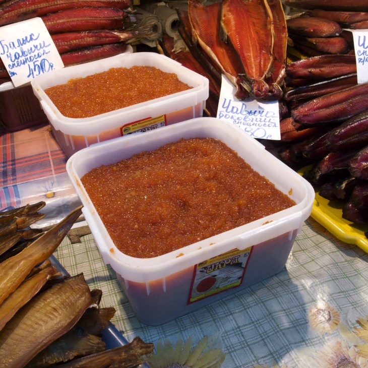 Containers of red salmon caviar displayed at a market stall with dried fish in the background.