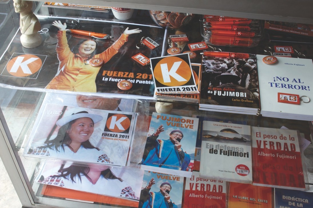 Display case of campaign materials featuring Fuerza 2011 posters, books about Alberto Fujimori, and promotional items with orange branding.
