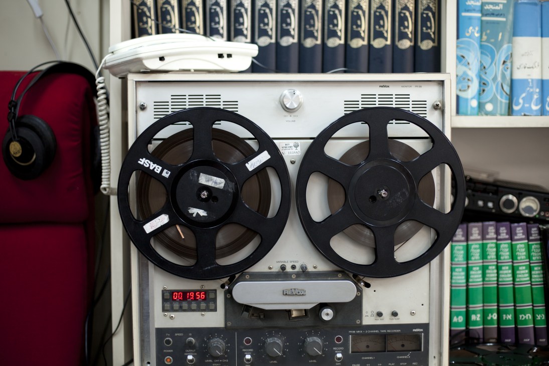 Reel-to-reel tape recorder with Hebrew and Farsi dictionaries on shelf behind it in radio studio.
