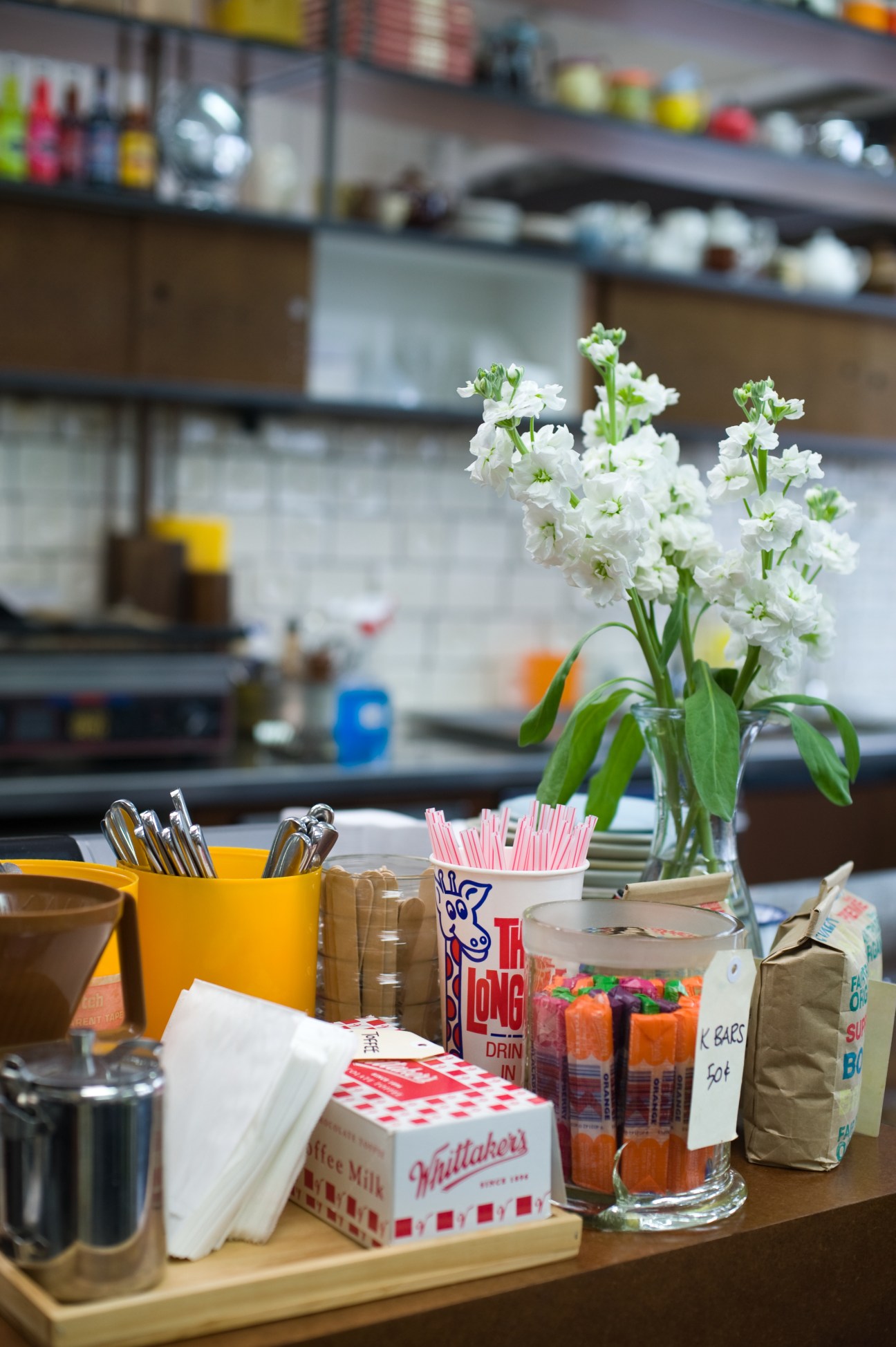 Café counter with white flowers in a vase, utensils, napkins, and condiments including L&P drink and Whittakers chocolate.