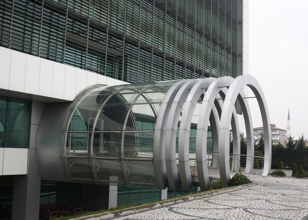 Modern curved glass and metal entrance structure attached to a contemporary building with horizontal louvers.