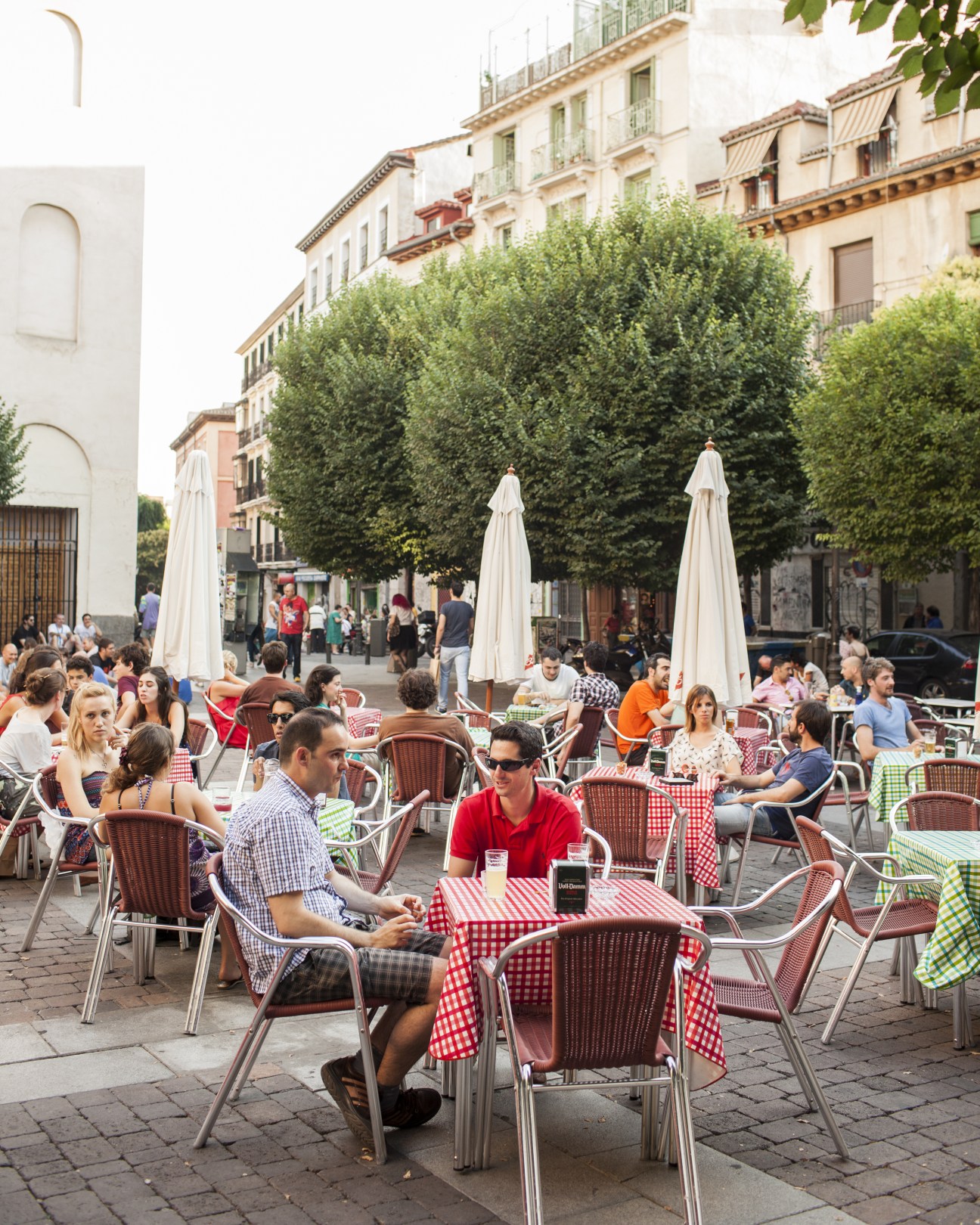Busy outdoor café terrace in Madrid's San Ildefonso Square with diners at checkered tables and white umbrellas.