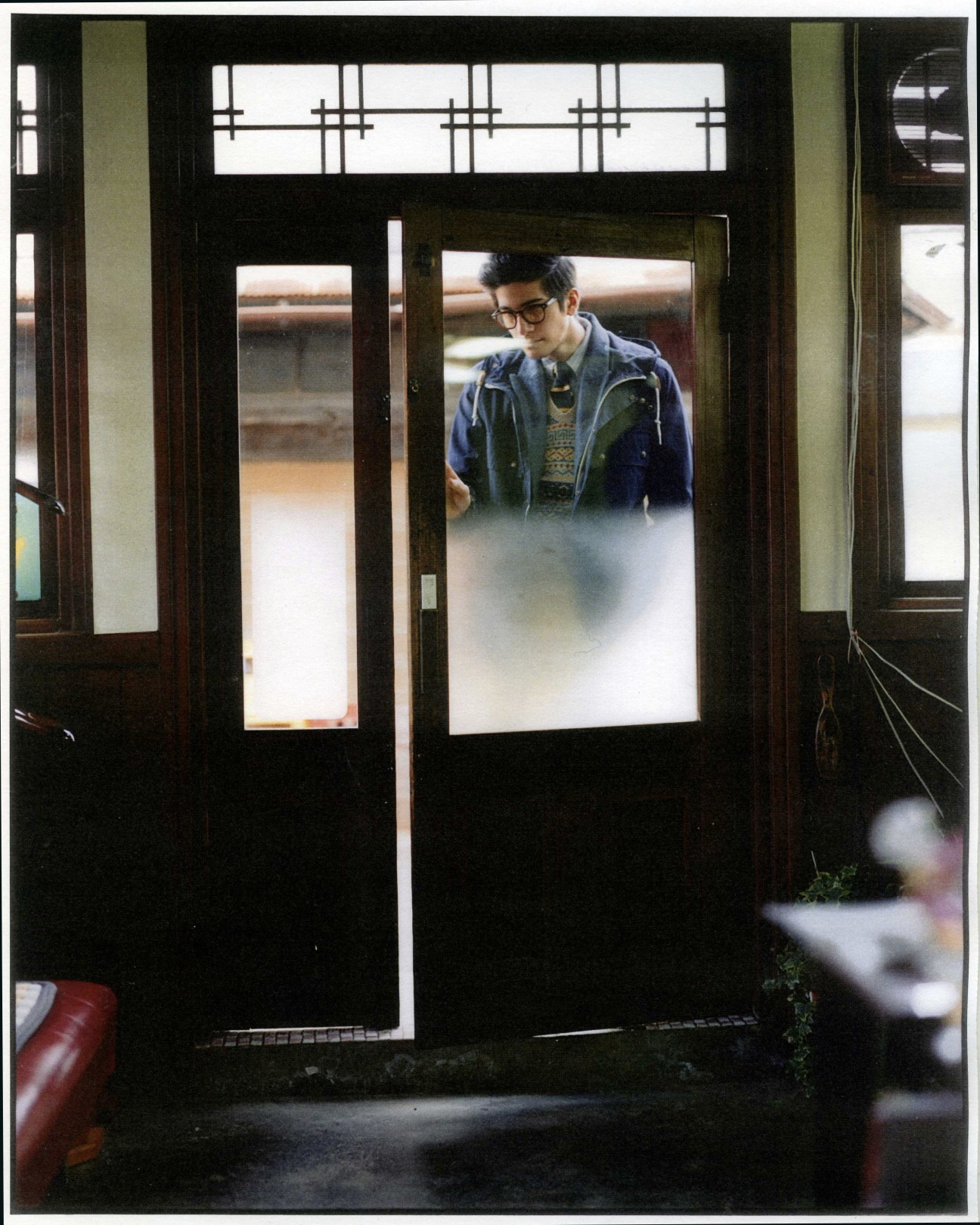 Man in glasses and layered clothing stands in doorway of traditional wooden-framed barber shop entrance.
