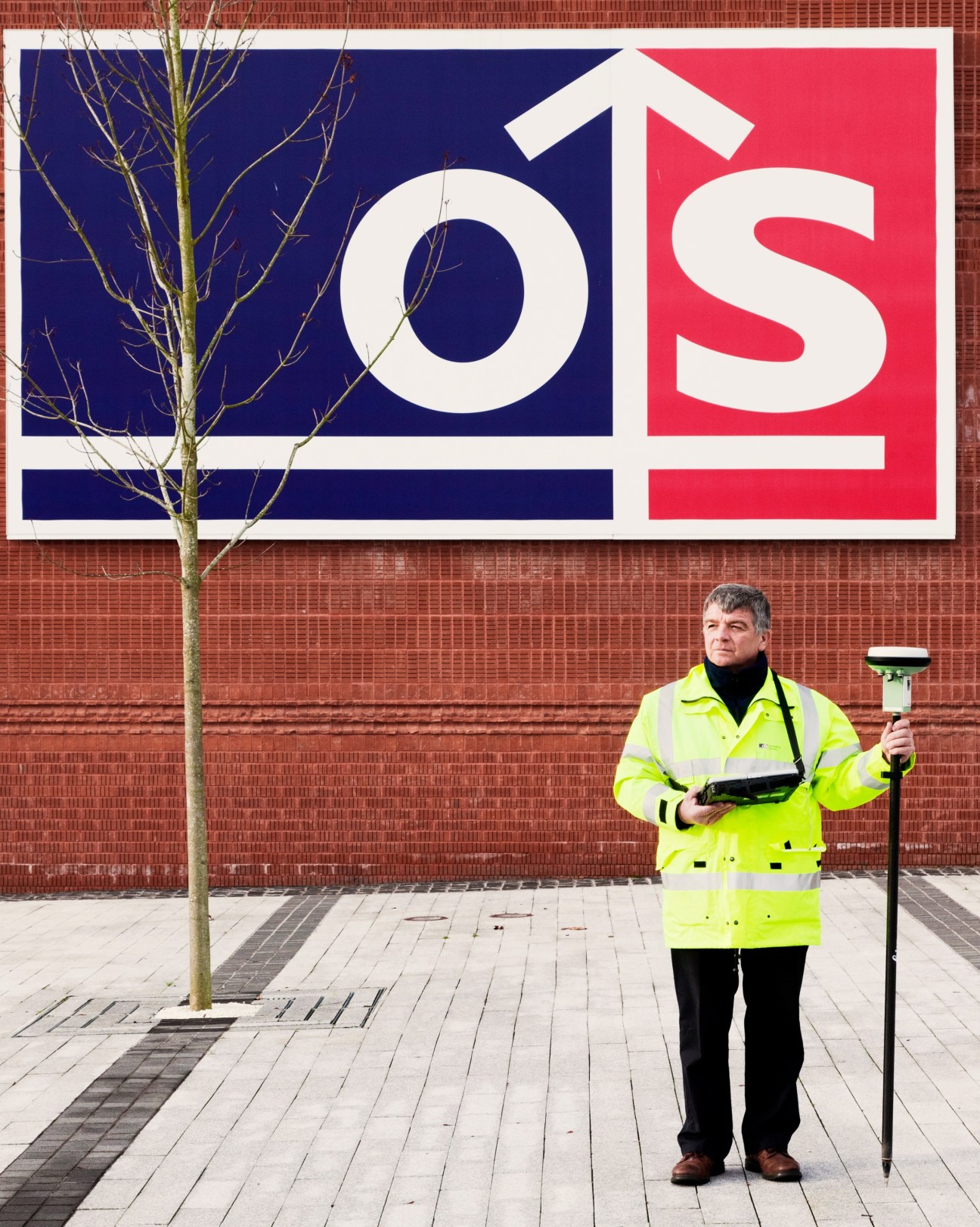 Nigel Vincent-Piper in high-visibility jacket holding surveying equipment outside Ordnance Survey headquarters.