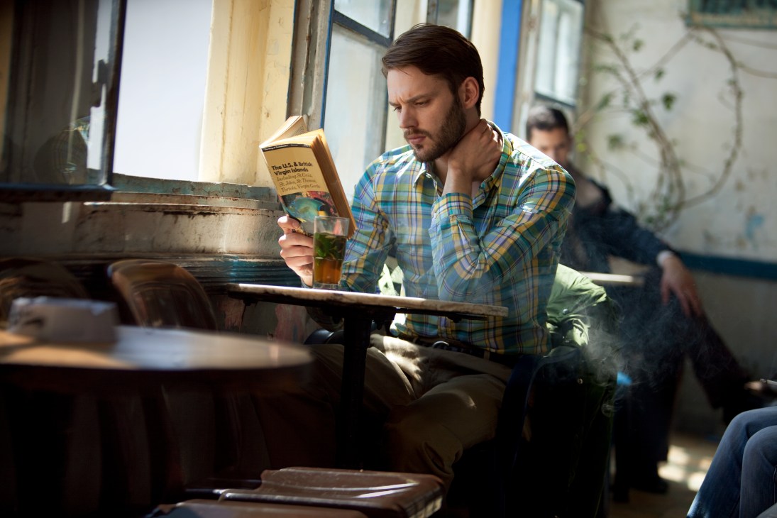 Man in plaid shirt reading at a table in a vintage café with worn wooden paneling and windows.