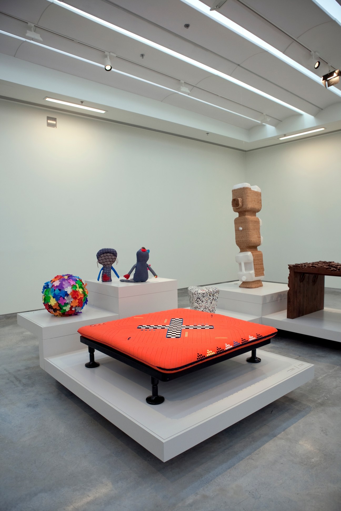 Contemporary art installation displaying colorful sculptures and objects on white pedestals in a minimalist gallery space.
