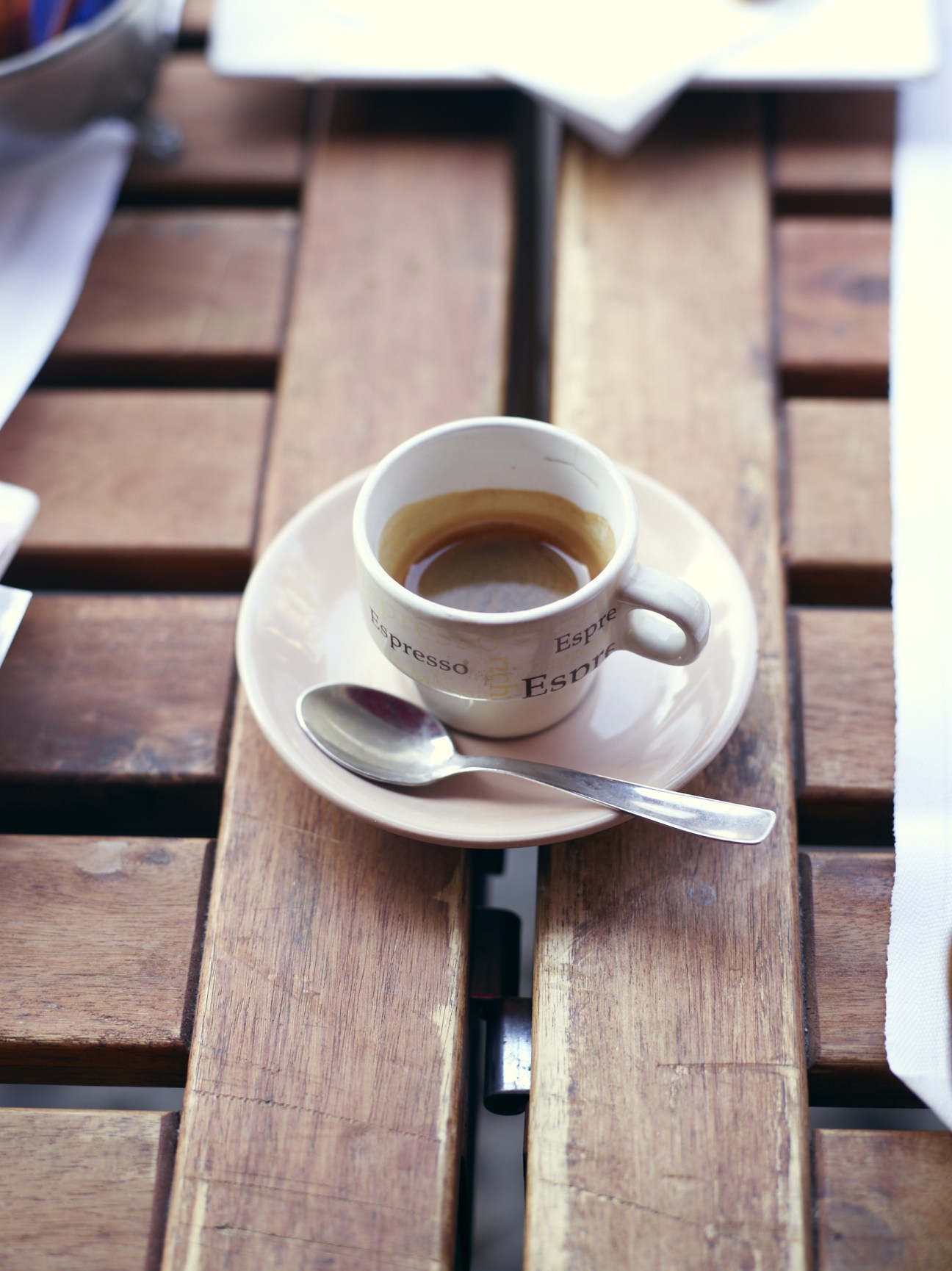 A cup of espresso with "Espresso" text printed on the white cup and saucer, placed on a wooden bench.