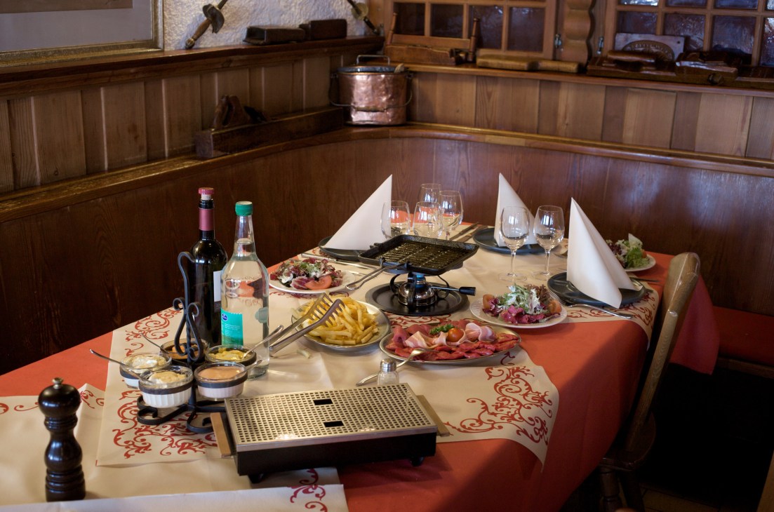 A wooden-paneled dining room table set with wine bottles, glasses, plates of sliced meat, fries, sauces, and a metal hot plate for cooking charbonnade.