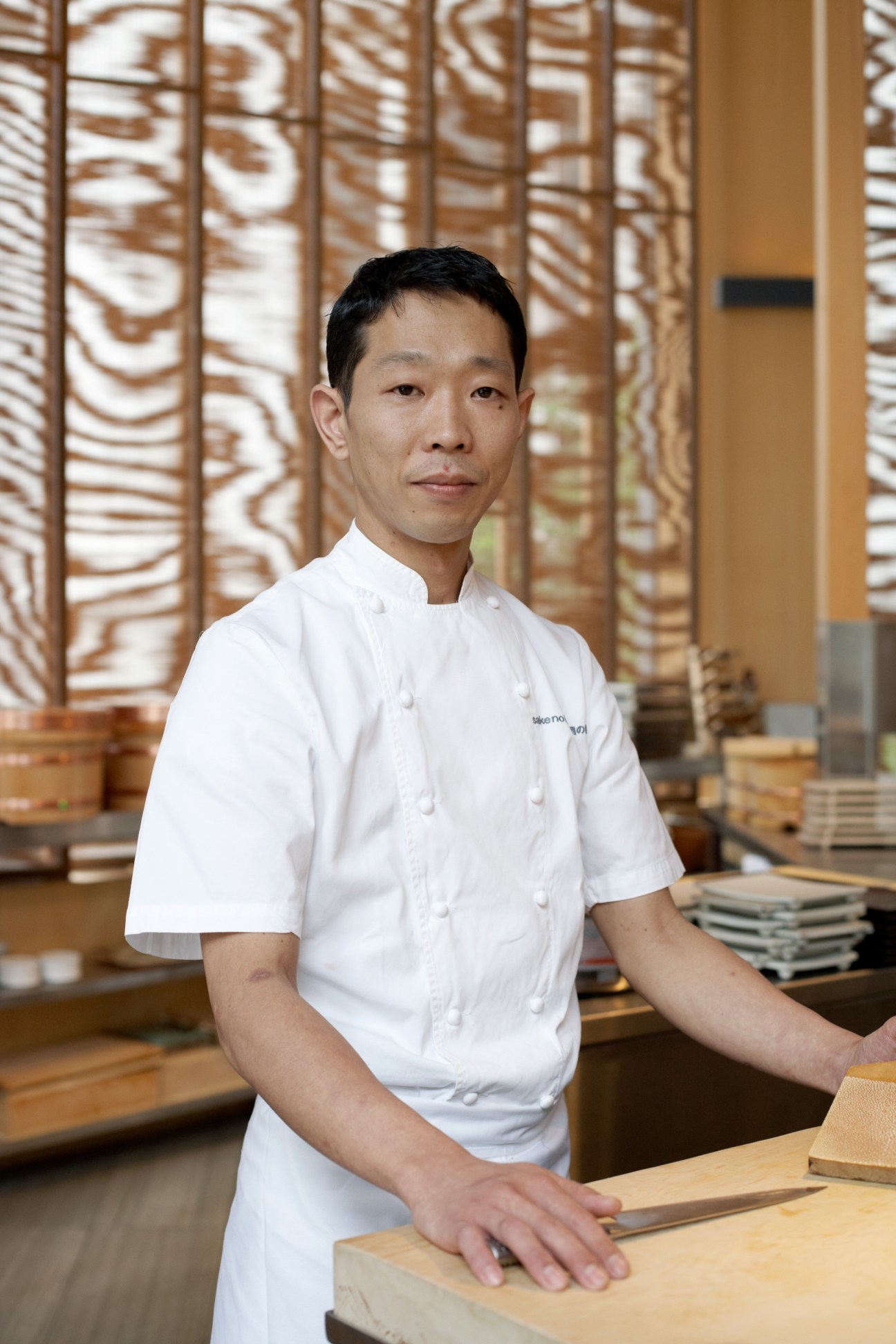 Chef in white uniform standing at counter with decorative wooden screens behind him at Saké no Hana restaurant.
