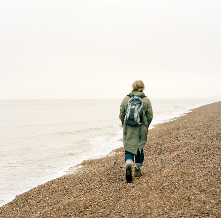 Woman jogging alone on a pebbly beach wearing a green jacket and backpack.