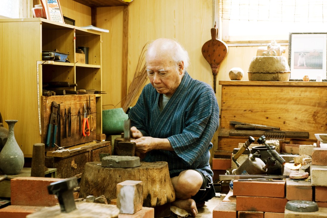 Ryukyu traditional goldsmith Kenjiro Matayoshi working at his wooden workbench surrounded by tools and materials.
