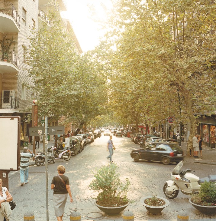 Tree-lined street in Naples with parked Vespas, pedestrians, and storefronts under dappled sunlight.