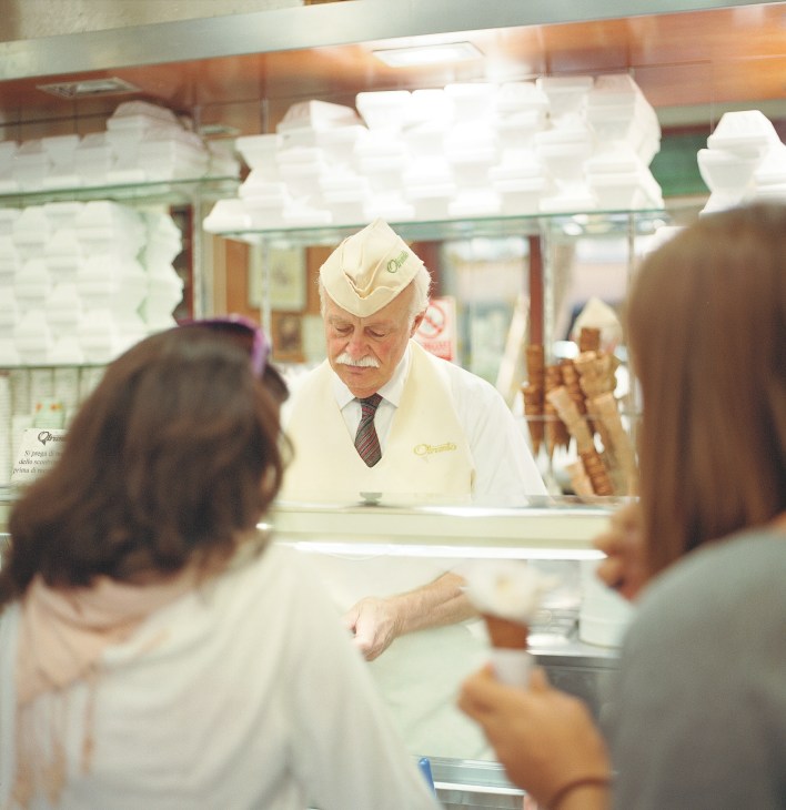Elderly gelato maker in white uniform and hat serves customers at Gelateria Otranto counter.