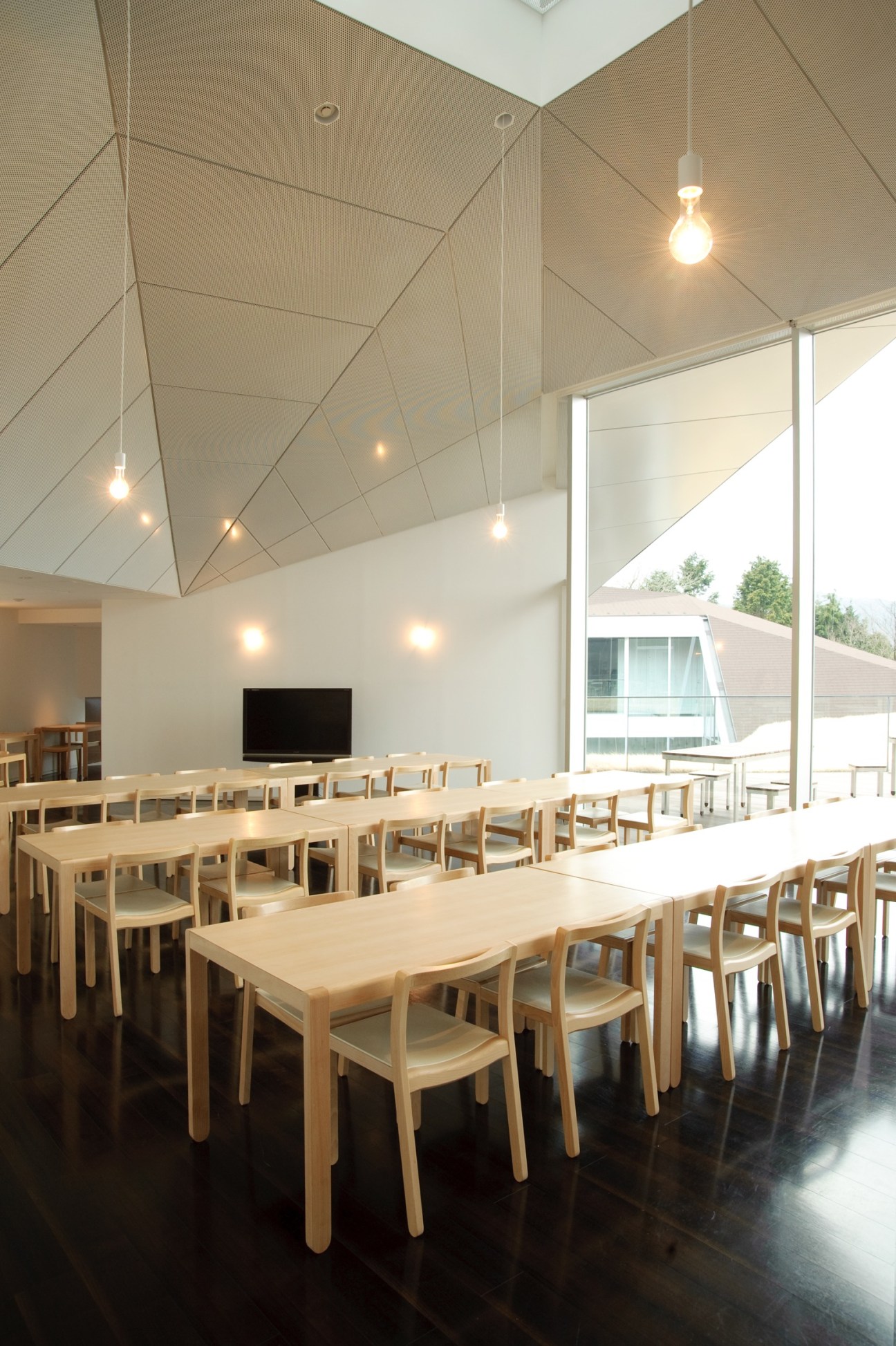 Top-floor restaurant with wooden tables, chairs, angular white ceiling with pendant lights, and floor-to-ceiling windows overlooking forest.