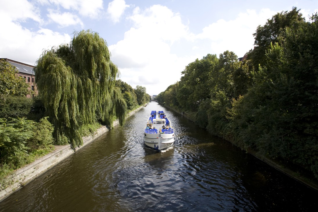 A tour boat with blue seating glides along the tree-lined Landwehr canal in Berlin's Neukölln district.