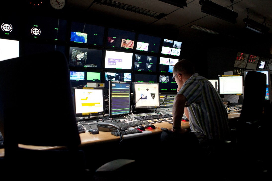 A technician works at a control room desk with multiple monitors displaying Channel 4 News branding and broadcast graphics.