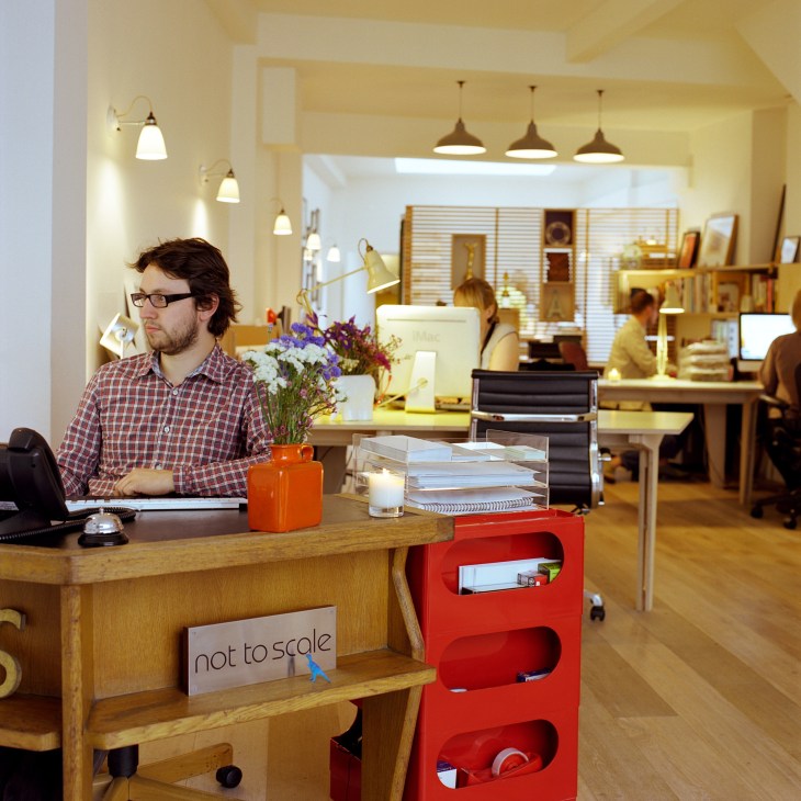 Not To Scale office interior with man at wooden desk, vintage pendant lights, flowers in orange vase, and colleagues working in background.