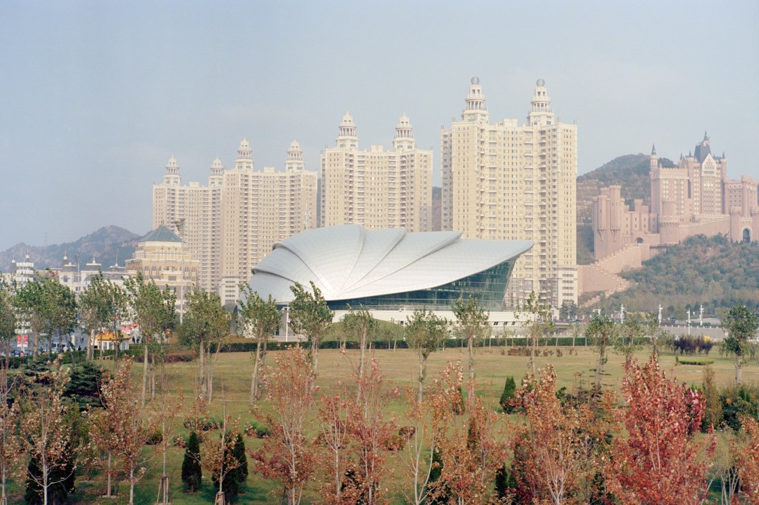 Dalian's Performing Arts Centre with white shell-like roof overlooks Xinghai Square with modern residential towers and wooded hills beyond.