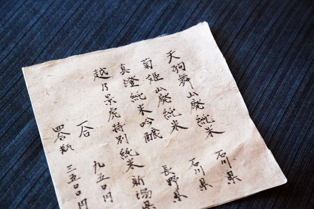 Handwritten menu in Japanese calligraphy on white paper against dark fabric.