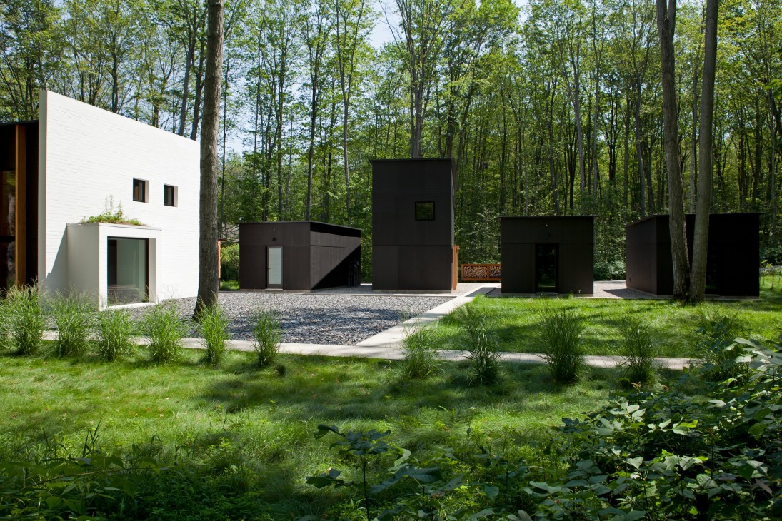 Modern outbuildings with white and black facades nestled among tall forest trees on a gravel courtyard.