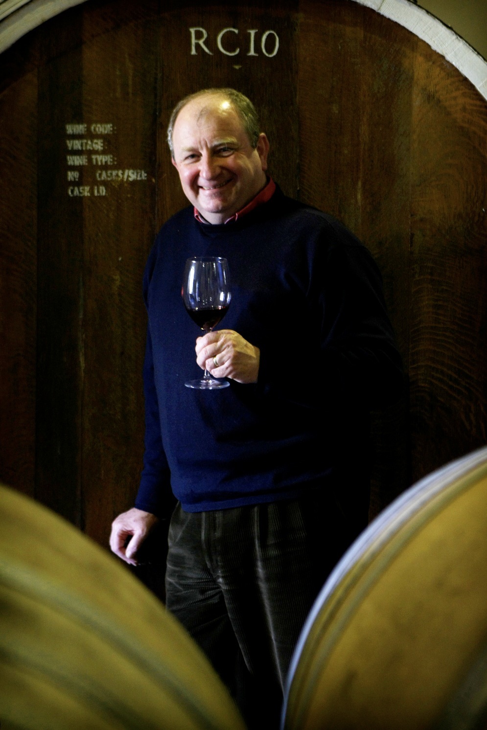 Winemaker John Cassegrain holds a glass of red wine in a cellar lined with wooden barrels.