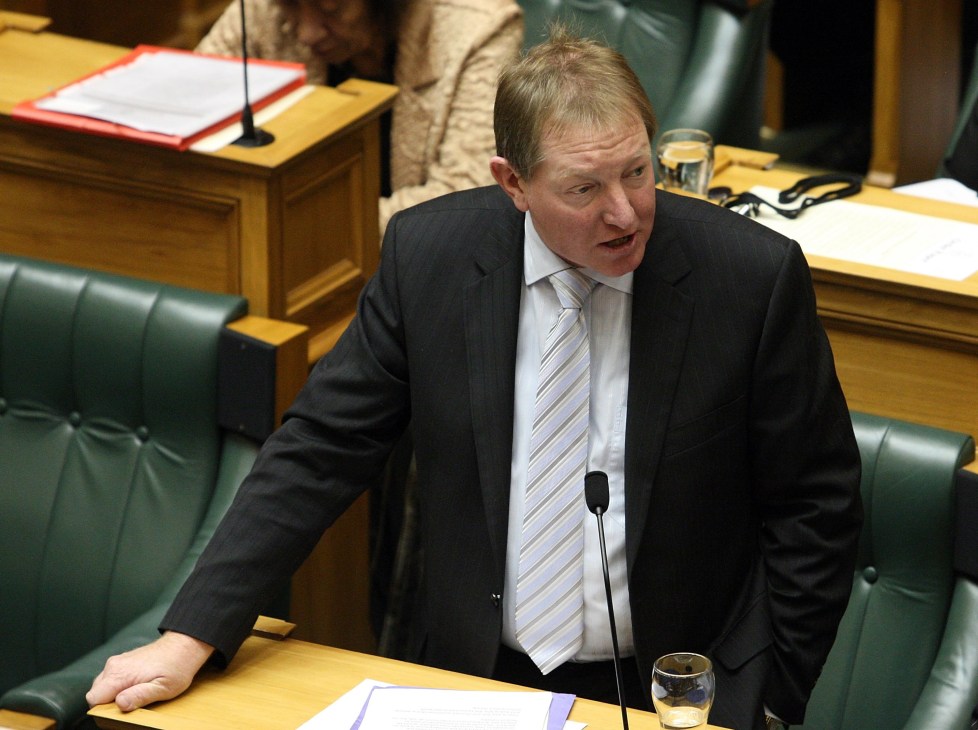 Man in dark suit and striped tie speaking at parliamentary dispatch box with microphone.