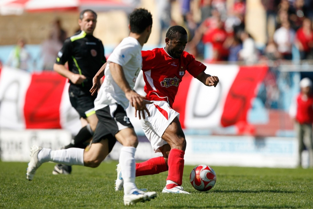 Football player in red jersey controlling ball during match with referee and opposing player in white.