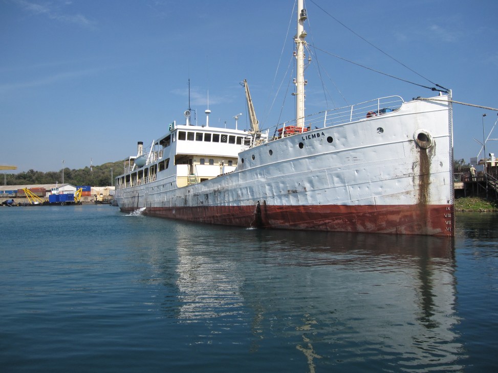 The MV Liemba, a historic white and red steamship, docked in calm blue water with industrial port facilities visible in the background.