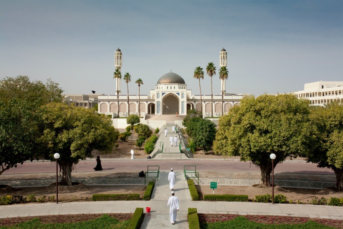 White mosque with golden dome and twin minarets on university campus surrounded by palm trees and manicured gardens.