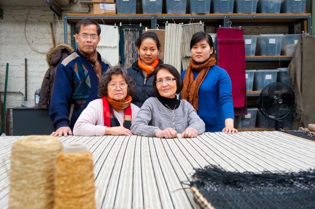 Cambodian artisans at Atelier Charles Schambourg workshop with woven leather samples displayed on shelves behind them.